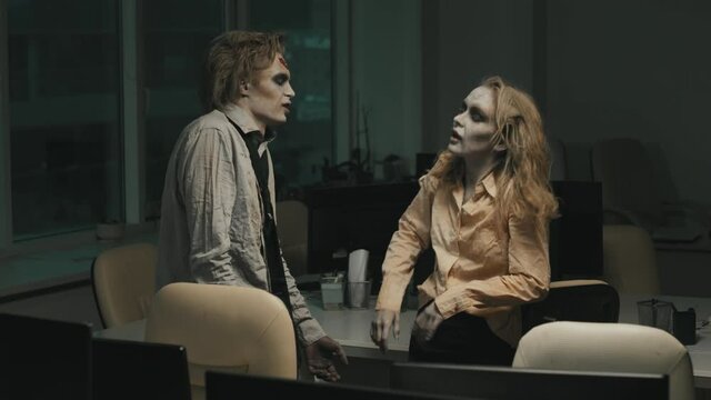 Medium shot with slowmo of grunting zombie man and woman with SFX makeup and torn dirty clothes having conversation in dark office