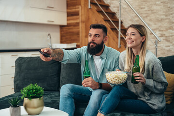 Young beautiful couple drinking beer, eating popcorn and watching sport game on a television at home © Zamrznuti tonovi