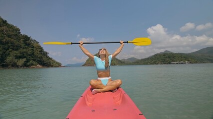 Attractive sportive blonde woman with sunglasses rows pink plastic kayak putting up paddle on sea against hills with wild jungles, blue sky.