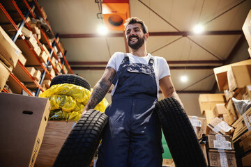 Low angle view of smiling hardworking blue collar worker in overalls carrying tires and walking in storage in import and export firm.