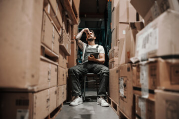 Young worried hardworking tattooed bearded blue collar worker in overalls sitting on the chair in...