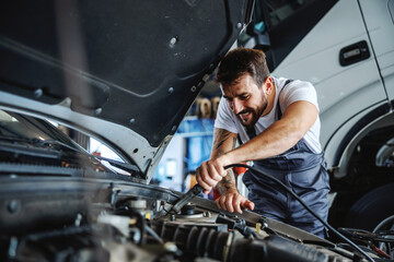 Hardworking dedicated bearded employee in overalls fixing motor. In background is truck. Manual...