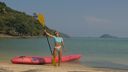 Pretty young sportswoman with sunglasses and swimsuit holds paddle posing near pink plastic canoe...