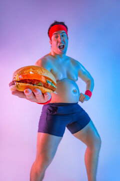 Fat man with gives away hamburger for lose weight and become a slim athlete. Not sporty male eating hamburger isolated on pink background.
