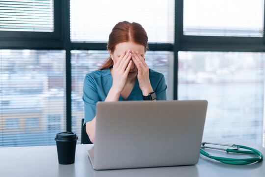 Overworked Young Female Doctor In Blue Medical Uniform Typing On Laptop And Rubbing Eyes Sitting At Desk On Background Of Window In Office Of Medic Clinic.