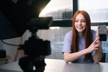 Close-up of cheerful young woman blogger talking about new phone using professional camera and softbox sitting at desk on background of window. Happy redhead female recording video vlog in office.