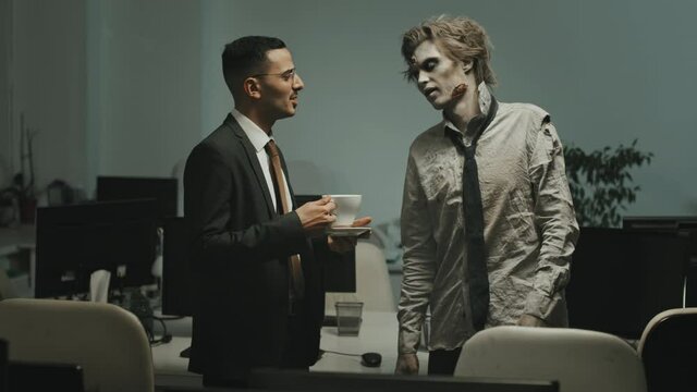 Medium shot of businessman in suit holding cup of coffee and talking to zombie man in torn shirt and necktie in office