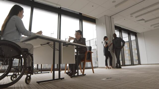Tracking left of disabled female HR manager sitting in wheelchair at desk in office and talking to young Asian man sitting in front of her
