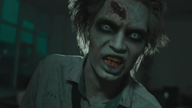 Close up shot of zombie man with SFX makeup with fake wounds and white contact lenses looking at camera and screaming in dark room with flashing lights