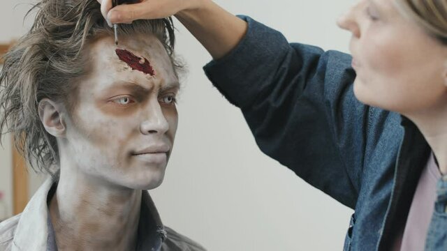 PAN shot of female SFX makeup artist creating fake wound on forehead of man with contact lenses and zombie makeup