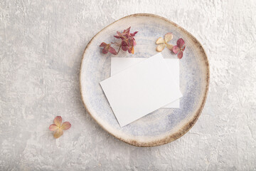 White paper invitation card, mockup with dried hydrangea flowers on ceramic plate and gray concrete background. flat lay, copy space.