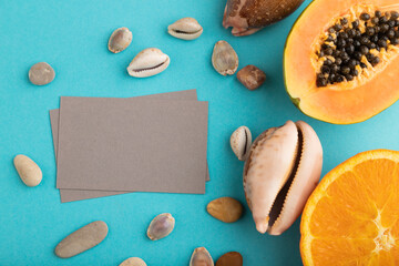 Gray paper business card with ripe cut papaya, orange, seashells on blue pastel pastel background. Top view, copy space, close up.