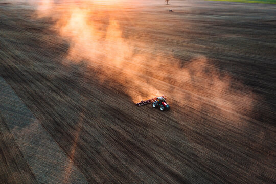 Aerial view of soil fields harrowing tractor and golden dust clouds behind him in countryside near Kaunas, Lithuania.