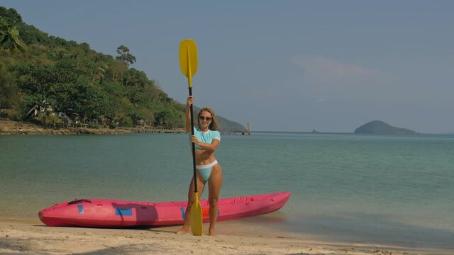 Pretty young sportswoman with sunglasses and swimsuit holds paddle posing near pink plastic canoe on sand beach at resort. Traveling to tropical countries. Attractive sexy girl posing near kayak.