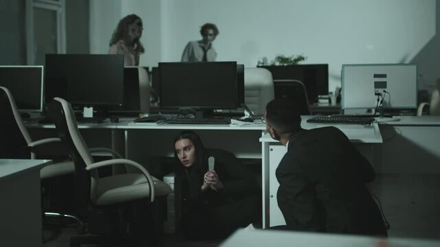 Handheld medium shot of businesswoman and businessman behind desks while zombies twitching and walking in office