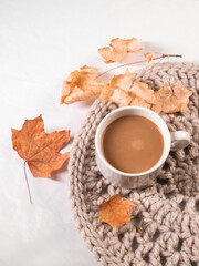 a cup of coffee with milk, autumn dry leaves, a white table, a knitted napkin stand
