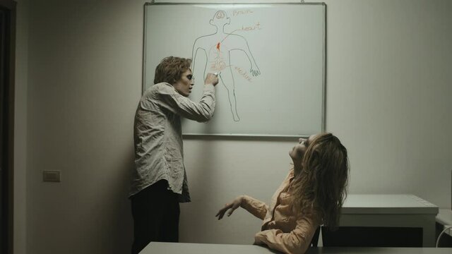 Medium shot of zombie man pointing at drawing of human on whiteboard and explaining different body parts to zombie woman