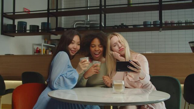Carefree mischievous pretty diverse multiethnic women in stylish outfit with coffee cups gathering at table, taking selfie shot on cellphone, making funny faces, expressing happiness and joy at cafe.