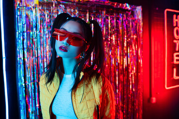 young asian woman in sunglasses looking at camera near red neon hotel sign