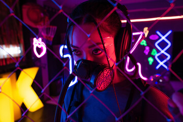 young asian woman in gas mask and wireless headphones near blurred metallic fence