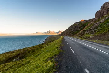 Papier Peint photo autocollant Atlantic Ocean Road Deserted road along a rugged coast of a fjord under clear sky  in summer. Some snow capped coastal mountains warmly lit by a setting sun are visible on horizon. Northern Iceland.
