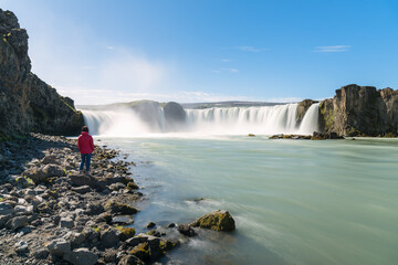 Woman in red jacket admiring a majestic waterfall while standing on rocks near the edge of a river on a clear summer day. Godafoss, Iceland.