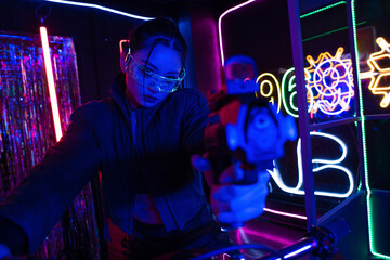 armed asian woman in sunglasses aiming with gun while riding motorcycle near neon sign