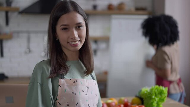 Slim gorgeous Korean woman with brown eyes and toothy smile posing in kitchen with African American friend at background. Portrait of beautiful confident lady in apron looking at camera smiling