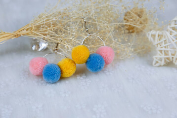 Colored pompon earrings in the shape of three furry balls.Earrings are next to rattan products, flower specimens and other ornaments