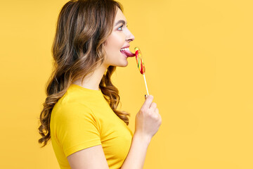 Profile portrait of young woman licking yummy colorful lollipop on yellow background