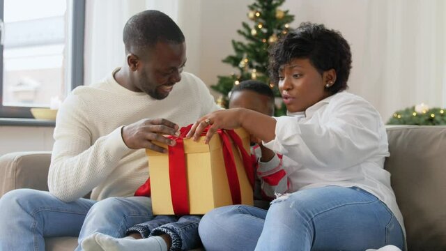 family, winter holidays and people concept - happy african american mother, father and baby son opening gift box with dinosaur toy at home on christmas