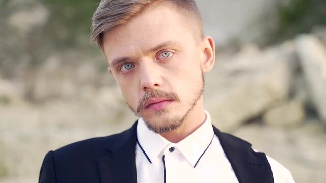 Close-up portrait of a young stylish fashion man looking at the camera. Outdoor. Trendy photogenic Fashionable male blond with a mustache and beard in a suit with blue eyes outside. Art fashion show