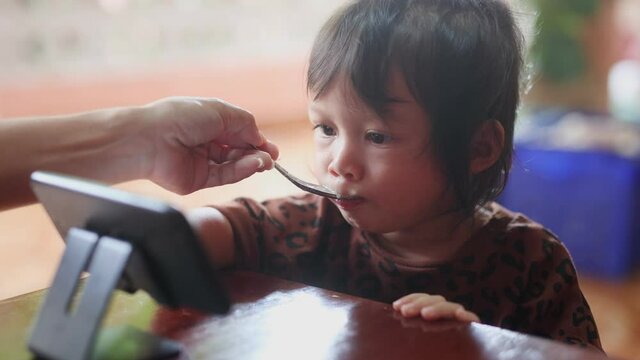 Asian woman, watched her smartphone with focus and attention while eating the food her mother fed. The concept of children is overly addicted to smartphones and is fed up with food.