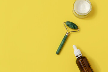 Above view of beauty roller made of jade stone with cream bottle and serum bottle on yellow background with space for text