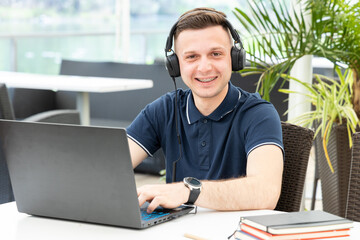 Student man watches a online webinar and listens to headphones while sitting at a table in a co...