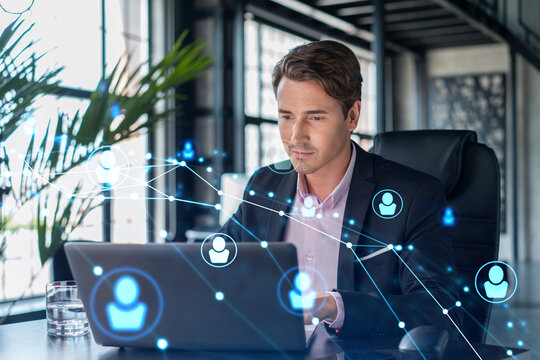 Handsome businessman in suit at workplace working with laptop to hire new employees for international business consulting. HR, social media hologram icons over office background