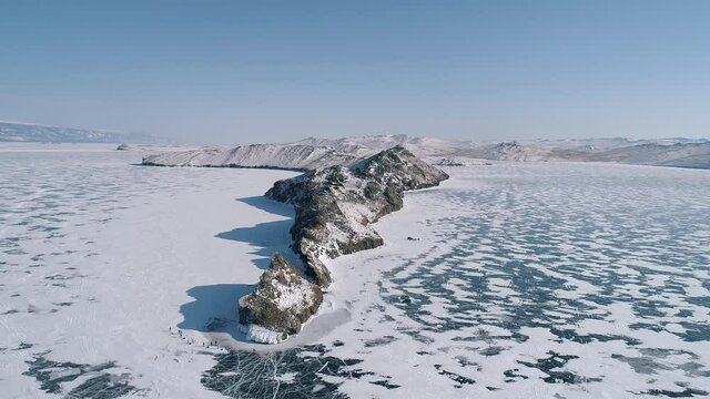 Aerial over the rocky cliff at lake Baikal. The blue snowy cracked ice of Baikal. Winter landscape of frozen Baikal. Groups of tourist walking around the cliff. Popular tourist spot