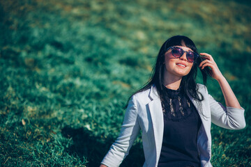 portrait of a life style brunette girl in sunglasses on the grass in the park in a white jacket