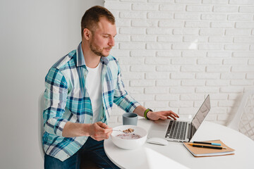 handsome smiling man in shirt sitting in kitchen at home at table working online on laptop