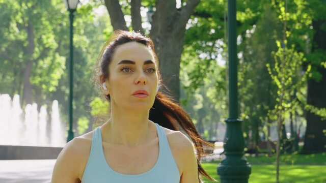 Young woman with ponytail wearing sports top and earphones jogging in green park, fountain on background. Tracking shot fit female exercising outside. Concept of healthy lifestyle
