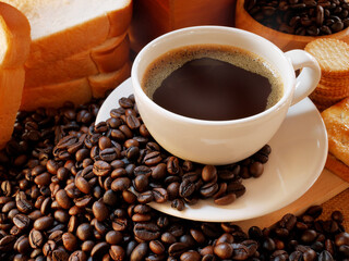 The hot aromatic coffee in a white cup is near some breads and some crackers and many coffee beans under its
