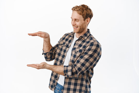 Image of caucasian redhead man smiling while looking at hands empty box, showing something, holding an object against white copy space, standing over white background
