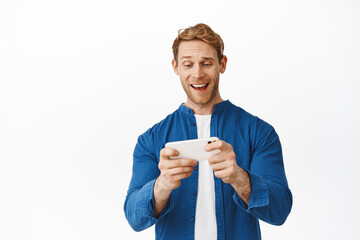 Happy redhead guy watching videos on smartphone, looking at mobile phone screen while playing video game app, smiling amused, standing over white background