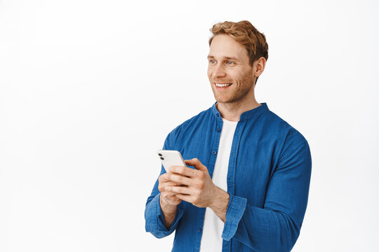 Handsome redhead man using smartphone application, smiling and looking left at advertisement logo with pleased face. Advertising of mobile phone app or shopping discounts, white background