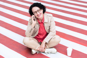 An adult woman with glasses with black hair sits on the sidewalk talking on the phone with a smile on her face..