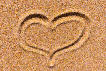 The symbol of the heart drawn on a sand. Background close up of a golden sunny sandy beach.