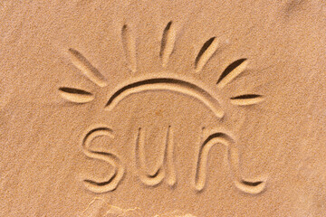 The text "Sun" is handwritten and sun rays are drawn on a golden sandy beach near the sea. Next to the text are waves of sand. Flat lay photo texture.