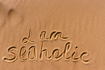 The text "I am seaholic" is handwritten on a golden sandy beach near the sea. Next to the text are waves of sand. At the top is copy space. Flat lay photo texture.