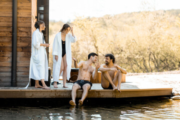 Company of friends rest on the terrace near the lake in the spa complex. Relax and fun time in wellness spa. Women in bathrobe walking on the terrace near their men.  - obrazy, fototapety, plakaty