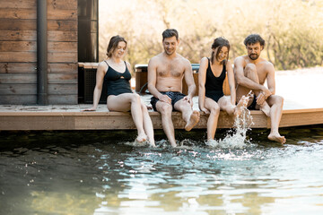 Company of friends sitting on the terrace and wetting their feet in the lake in the spa complex. Relax and fun time in wellness spa.